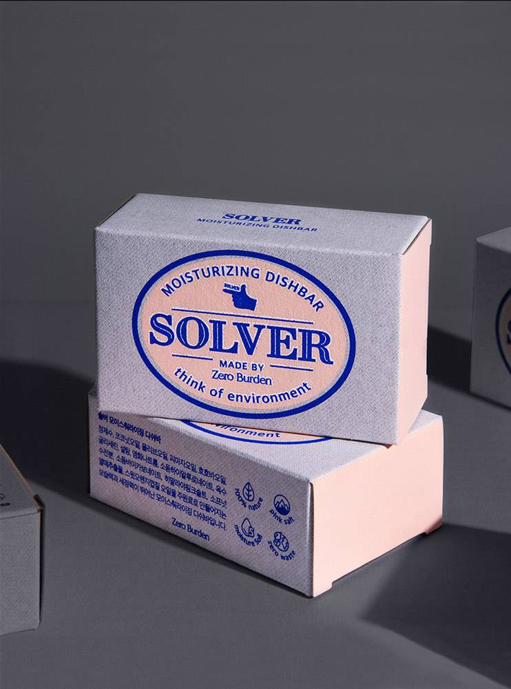 Solver 디쉬바 패키지가 바닥에 놓여 있는 장면으로, 작은 박스 두 개가 겹쳐진 상태로 정렬되어 있음. 패키지의 앞면에는 'Solver'라는 제품명이 선명하게 인쇄되어 있으며, 자연스러운 핑크색과 파란색의 조화가 브랜드의 친환경적 이미지를 강조하고 있음. 패키지의 깔끔한 디자인과 자연스러운 질감이 돋보임