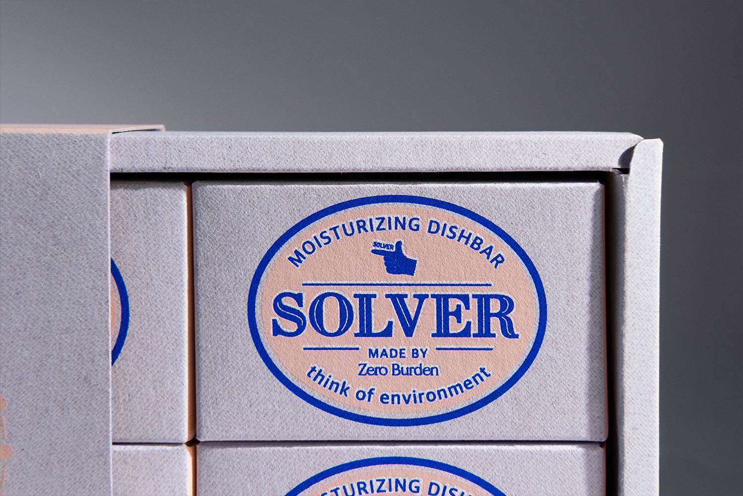 Solver 디쉬바 패키지의 일부를 클로즈업하여 촬영한 장면으로, 작은 박스 두 개가 겹쳐진 상태로 정렬되어 있음. 상단 박스에는 제품명이 파란색으로 선명하게 인쇄되어 있으며, 핑크색 배경과 함께 브랜드의 자연친화적이고 간결한 이미지를 잘 보여줌. 패키지 표면의 질감이 고급스럽고 자연스러움을 강조하고 있음