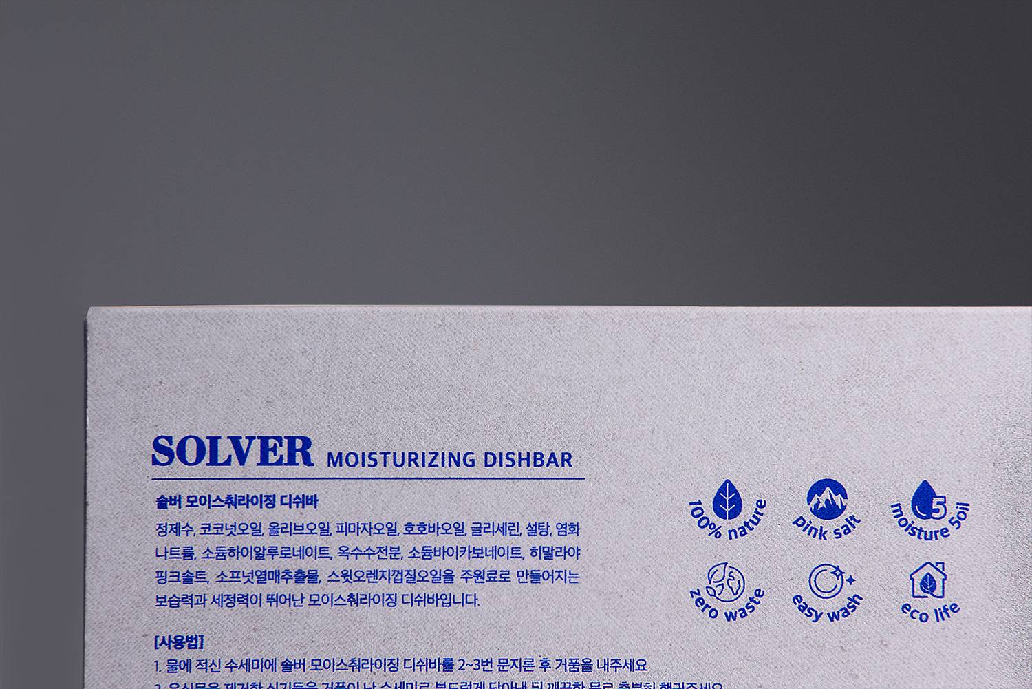 Solver 디쉬바 패키지의 뒷면을 클로즈업하여 촬영한 장면으로, 제품의 성분과 사용법이 파란색 텍스트로 인쇄되어 있음. 주요 성분으로는 히말라야 핑크 솔트, 코코넛 오일 등이 있으며, 각 성분의 특성과 제품의 장점을 아이콘과 함께 설명하고 있음. 자연 친화적이며, 환경을 고려한 포장 디자인이 돋보임