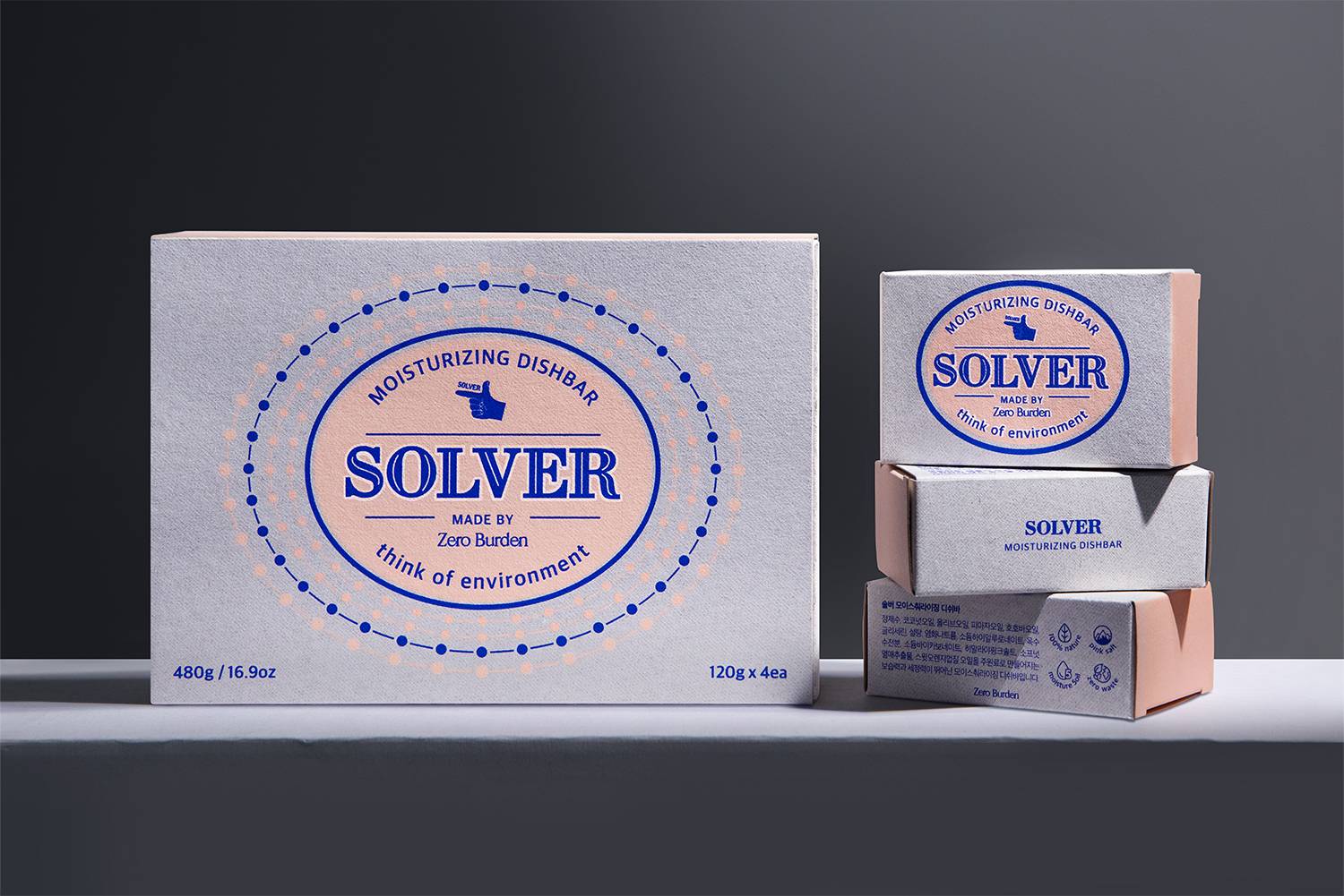 Solver 디쉬바 패키지 디자인이 담긴 사진. 중앙에는 핑크색 배경에 파란색 텍스트로 'Solver'라는 제품명이 강조되어 있음. 왼쪽 큰 상자와 오른쪽 쌓여있는 작은 상자들이 함께 보여지고 있으며, 패키지 표면은 무광 크라프트 용지로 만들어져 자연스러운 질감을 강조하고 있음
